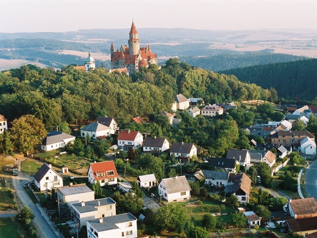 Panoramatický letecký pohled na hrad Bouzov, středověký hrad s červenou střechou a vysokou věží, stojící na zalesněném kopci. V popředí je vidět malebná vesnice s bílými domy a zahradami. V pozadí se rozprostírá zvlněná krajina s kopci a lesy.