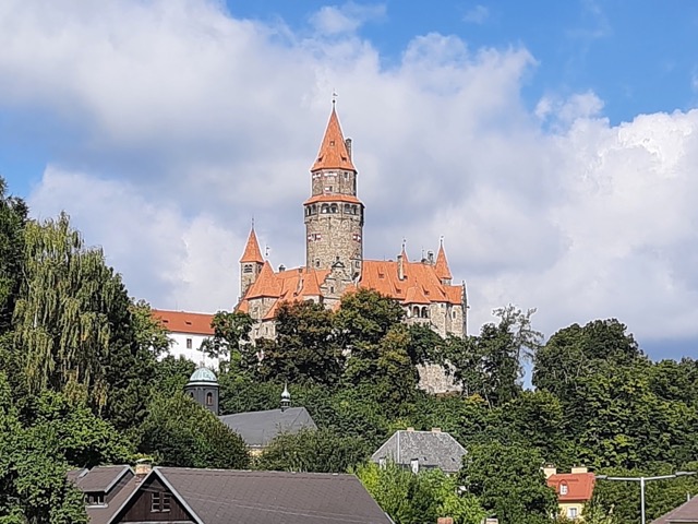 Pohled na hrad Bouzov zespodu, který se tyčí nad zelenými stromy. Hrad má kamenné zdi, červenou střechu a vysokou věž. V popředí jsou vidět střechy domů a listnaté stromy. Obloha je modrá s bílými mraky.