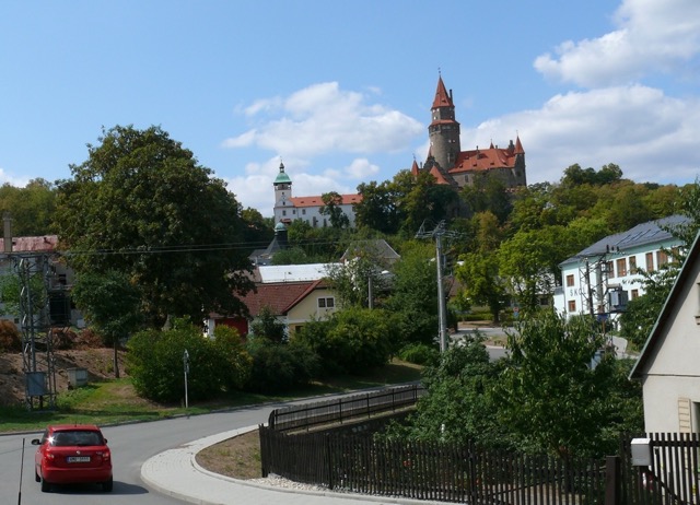 Pohled na hrad Bouzov z vyvýšeného místa, s červeným autem v popředí. Hrad má kamenné zdi, červenou střechu a vysokou věž. Je obklopený zelenými stromy a domy s červenými střechami. V pozadí je modrá obloha s bílými mraky. V popředí je vidět silnice s červeným autem a plotem.